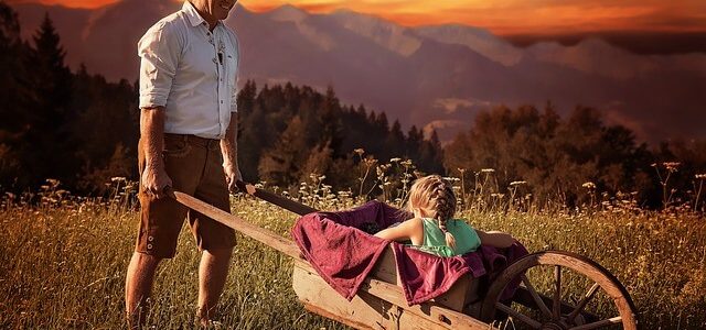 Jak spędzić wakacje z dzieckiem? Pomysły na urlop z maluchem Mężczyzna wiezie dziecko na taczce, w oddali widać szczyty gór